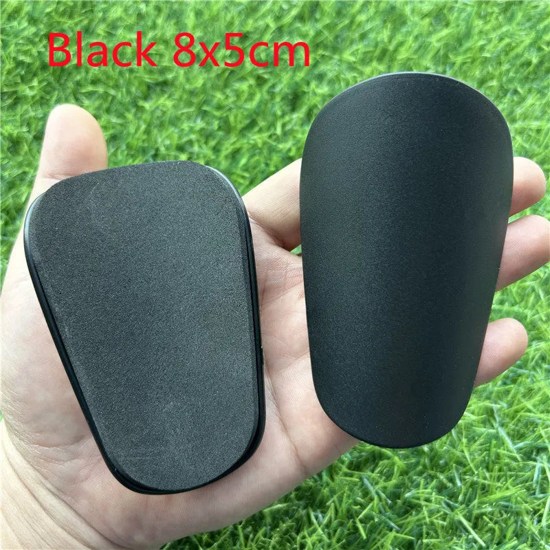 Extra small black and white plastic football shin guards for junior players, shown at 8x5 cm size.