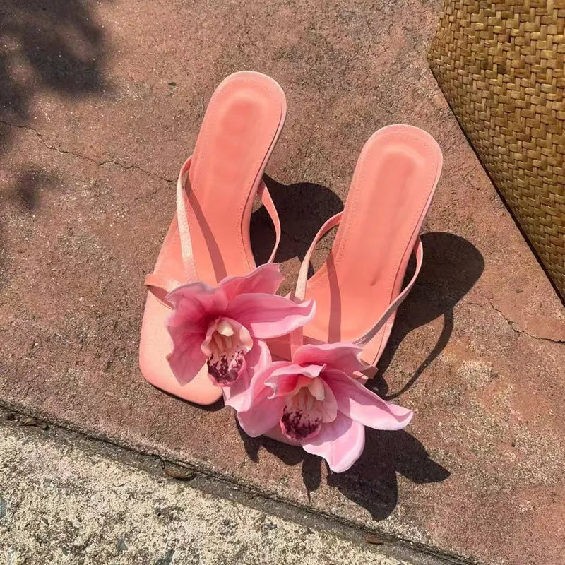 Pink women's sandals with a large artificial Phalaenopsis flower on the satin upper, a mid-heel, and a square toe.