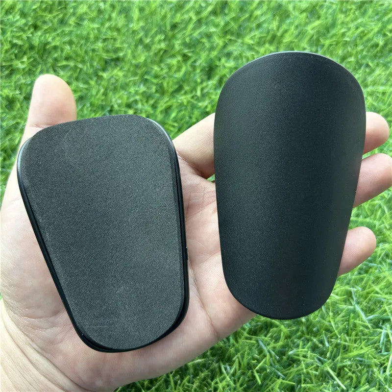 Extra small black and white plastic football shin guards for junior players, shown at 8x5 cm size.