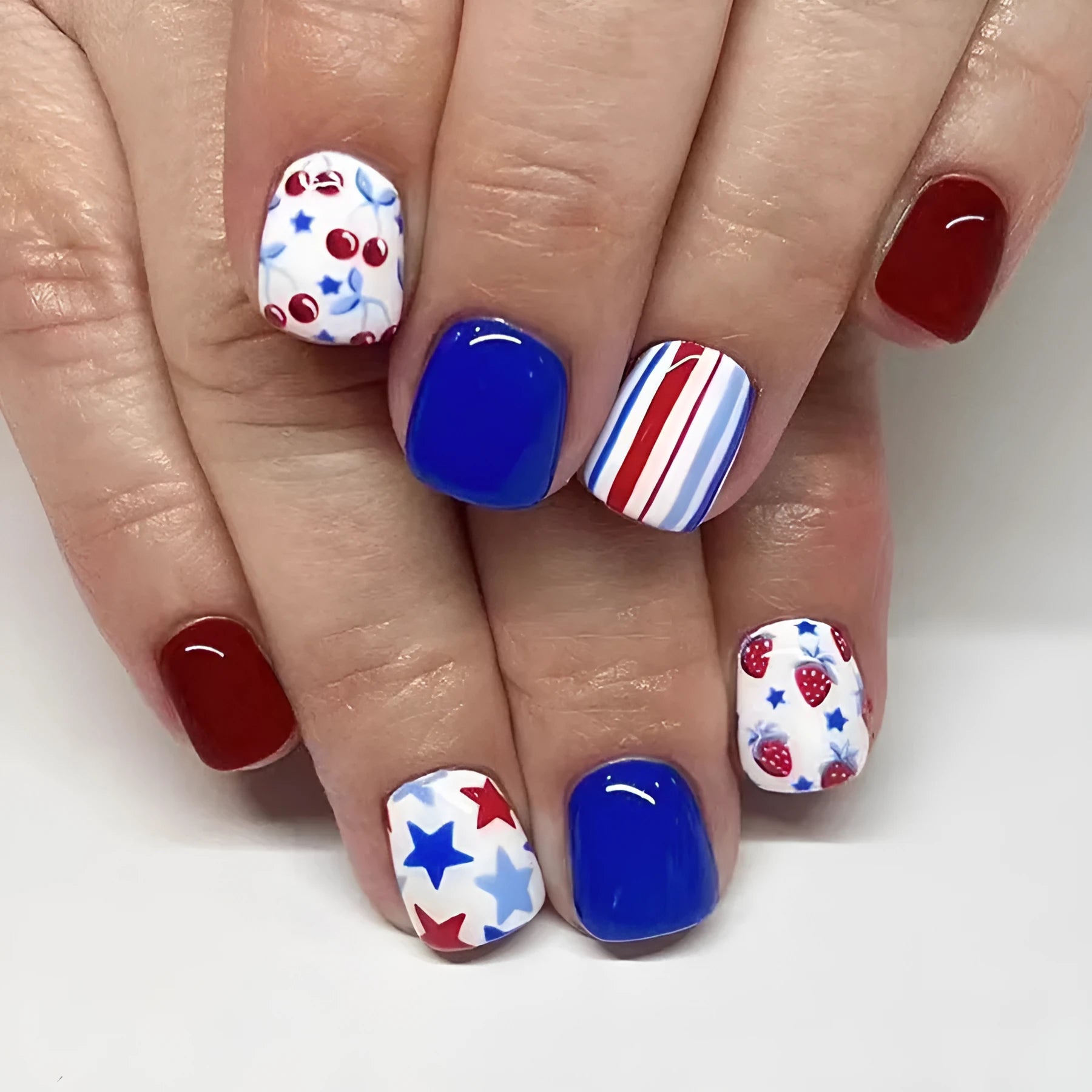 Short glossy false nails with strawberry and cherry pattern on red and blue background.