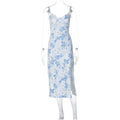 A woman wearing a long blue dress with a lace floral print, a side slit and thin suspender straps.