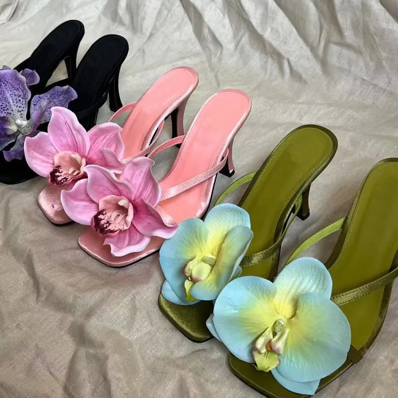 Pink women's sandals with a large artificial Phalaenopsis flower on the satin upper, a mid-heel, and a square toe.