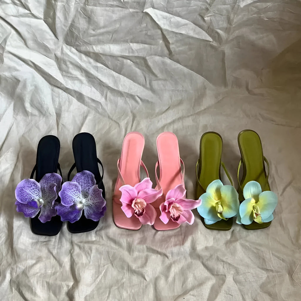 Pink women's sandals with a large artificial Phalaenopsis flower on the satin upper, a mid-heel, and a square toe.