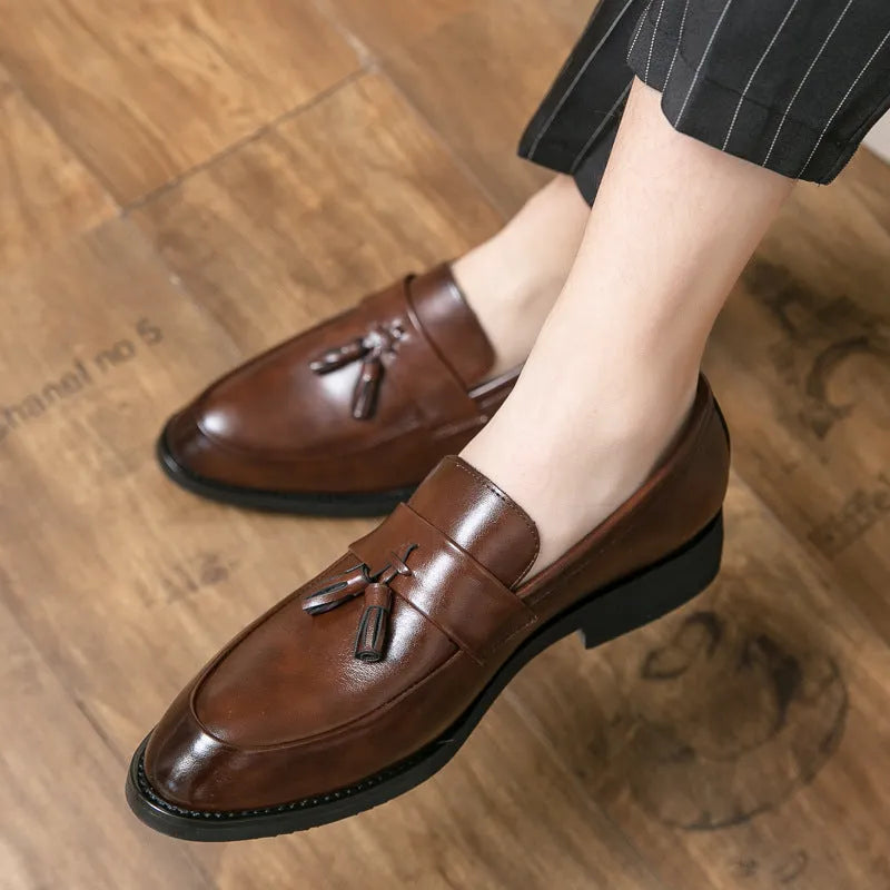 Men's black tassel loafers with a slip-on design and rubber sole on a plain background.