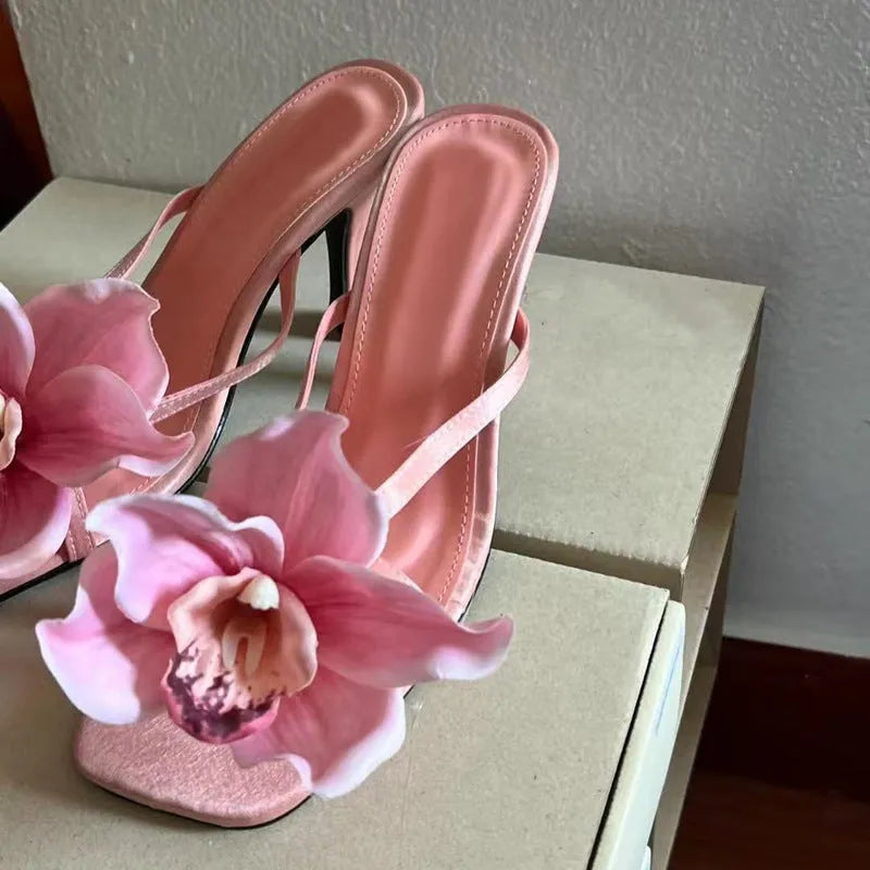 Pink women's sandals with a large artificial Phalaenopsis flower on the satin upper, a mid-heel, and a square toe.