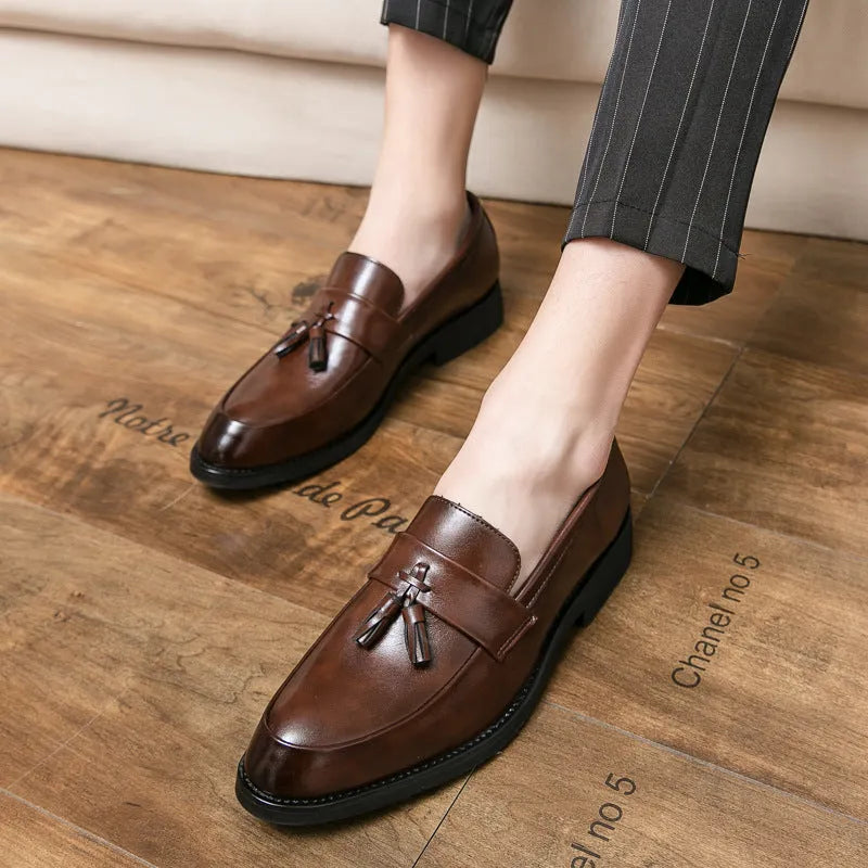 Men's black tassel loafers with a slip-on design and rubber sole on a plain background.