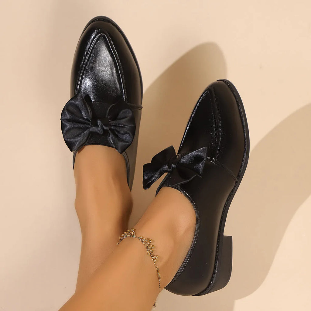 Women's brown slip-on flat shoes with a large bow decoration on the pointed toe.